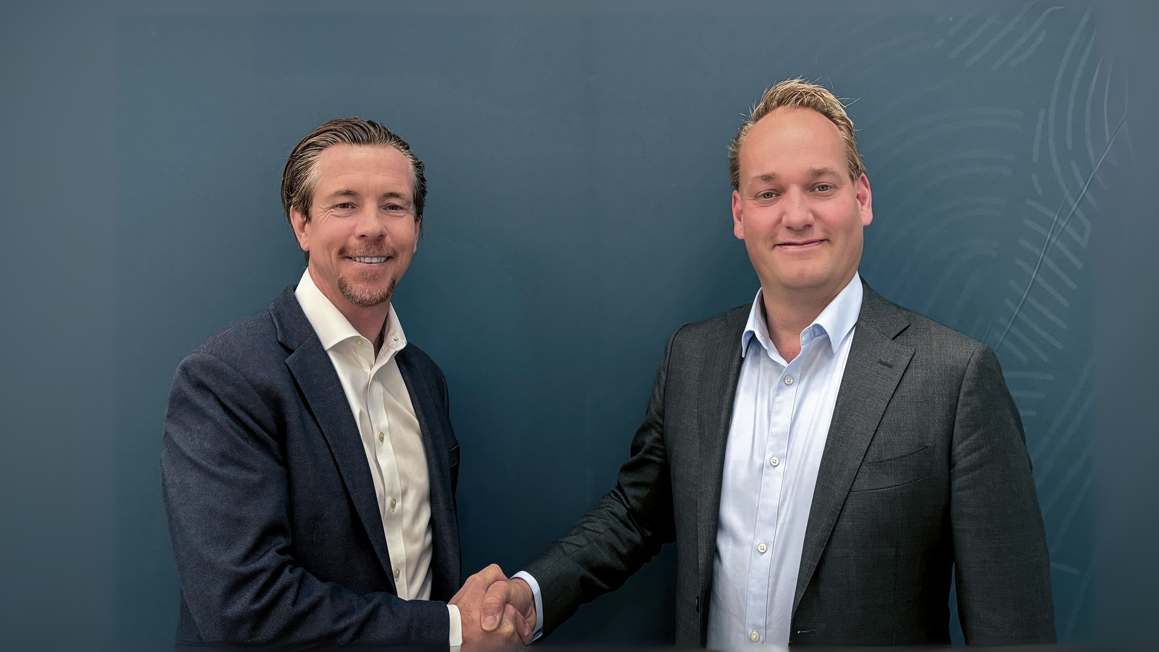 Two men in suits shaking hands in front of a blue background, smiling at the camera.